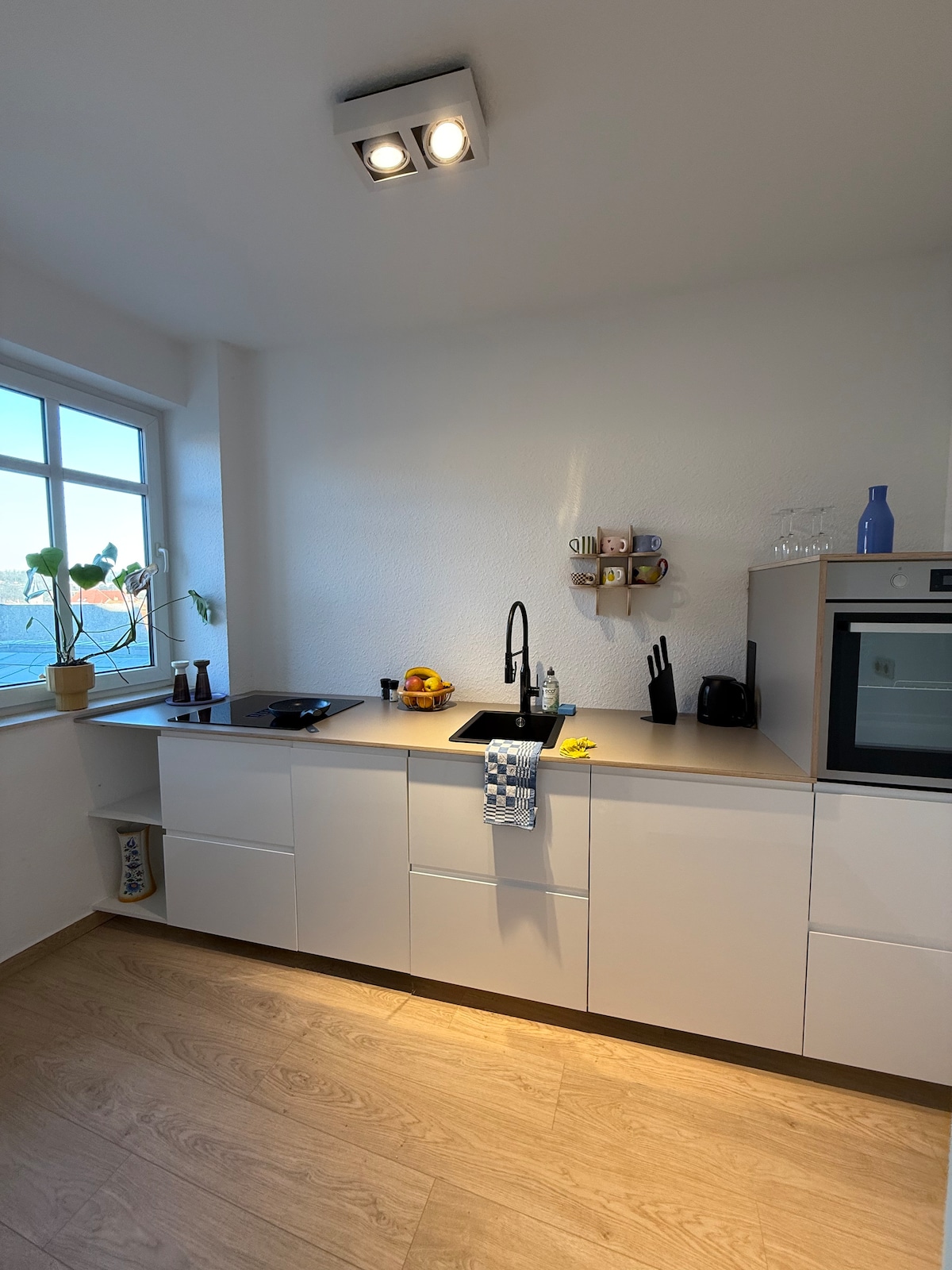The kitchen features modern cabinetry with a smooth countertop and built-in appliances. A sleek black faucet sits above the sink, accompanied by a coffee maker and various kitchen utensils. Natural light enters through a window, illuminating the organized space.