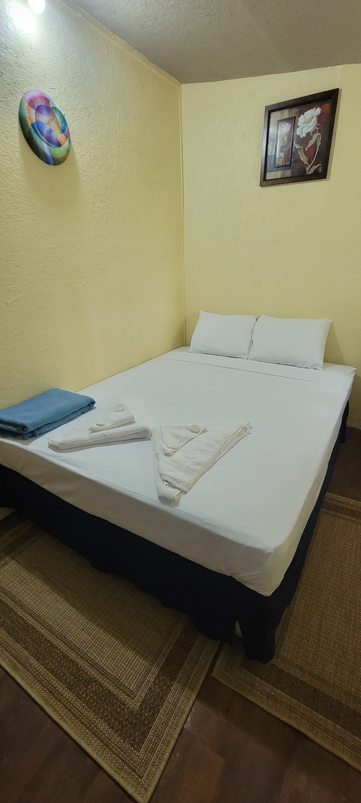 A cozy sleeping area features a neatly made bed with white bedding and two accent pillows. Towels are arranged on the bed, complemented by a blue blanket nearby. The warm yellow walls create a calming environment, and artwork is displayed above the bed.