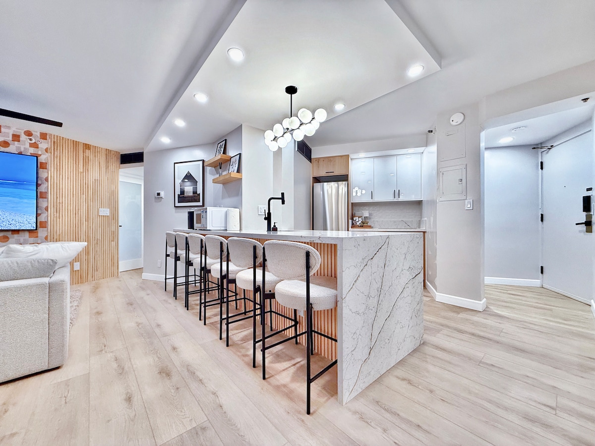 A modern kitchen with a large island features seating for six. The island is complemented by sleek cabinetry and stainless steel appliances, including a refrigerator and built-in oven. Bright, overhead lighting enhances the open, airy atmosphere of the space.