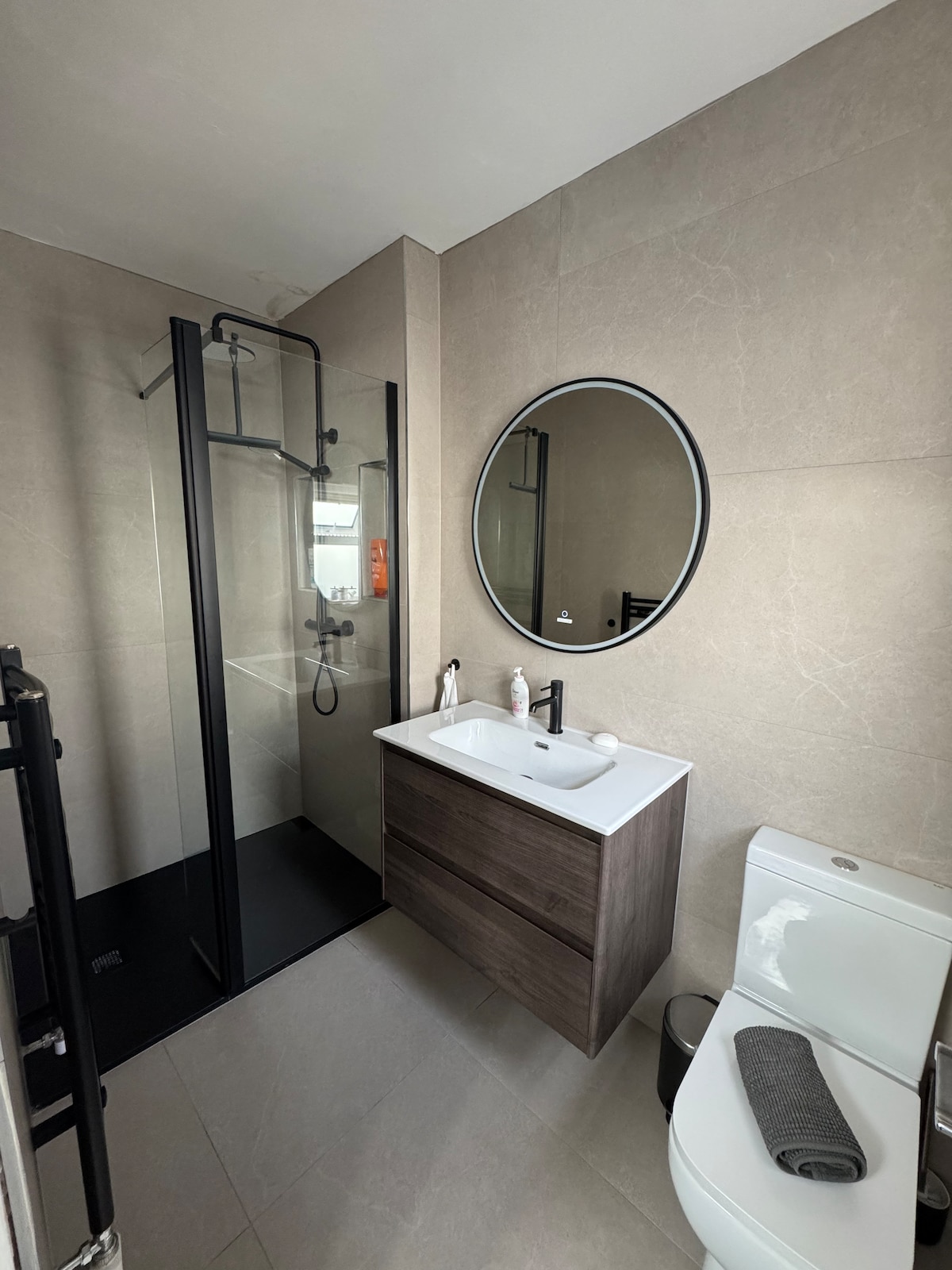 A modern bathroom features a glass walk-in shower with a sleek black frame. A circular mirror is mounted above a compact wooden vanity with minimalistic design. Light-colored stone walls enhance the space, while a neatly rolled gray towel rests on the bathroom’s white toilet.