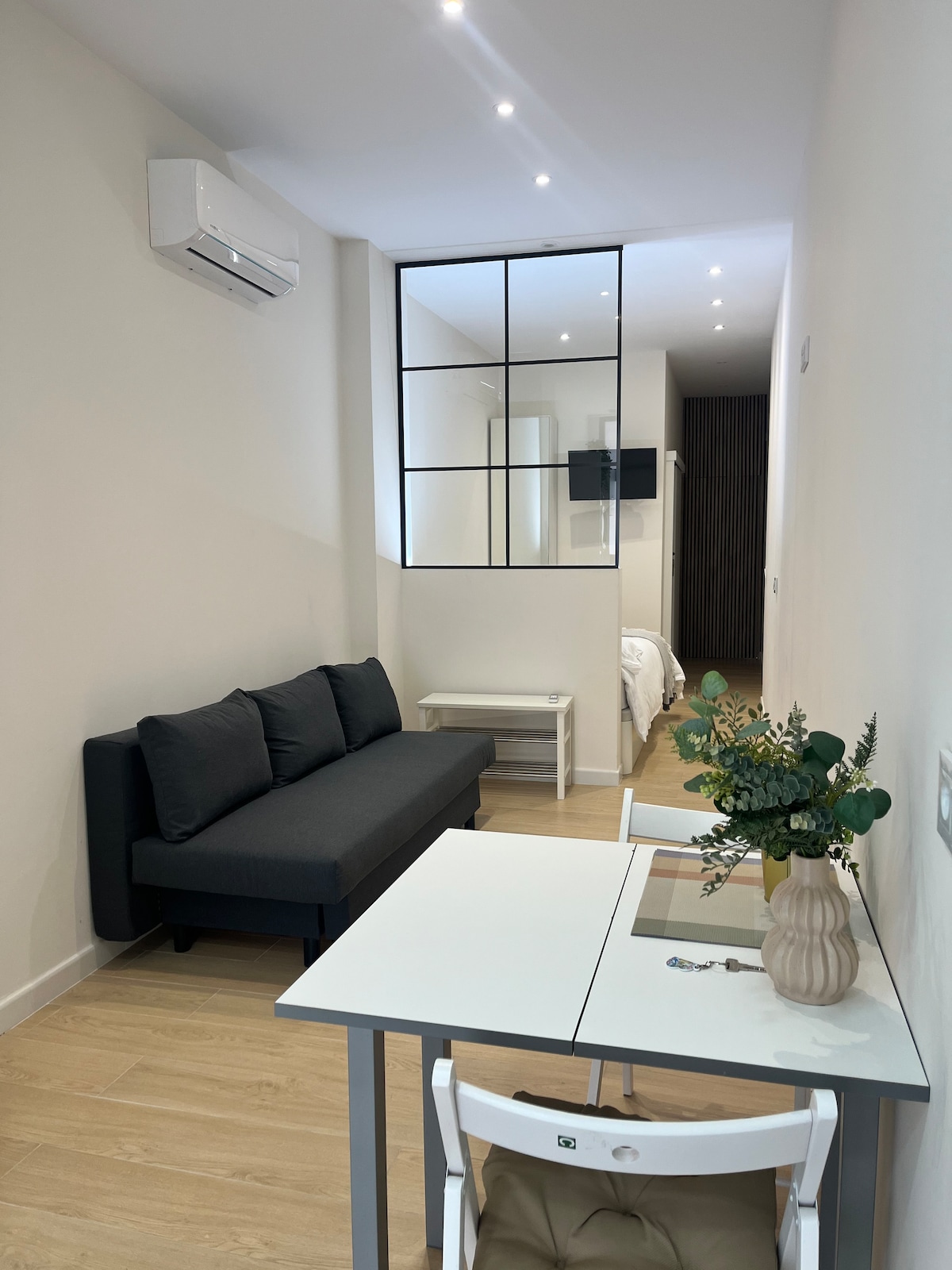 A well-lit living space features a dark sofa and a small dining area with a white table and chairs. A stylish vase with greenery adds a touch of decor. Reflections from a glass partition provide a view into a separate sleeping area, enhancing the sense of openness.