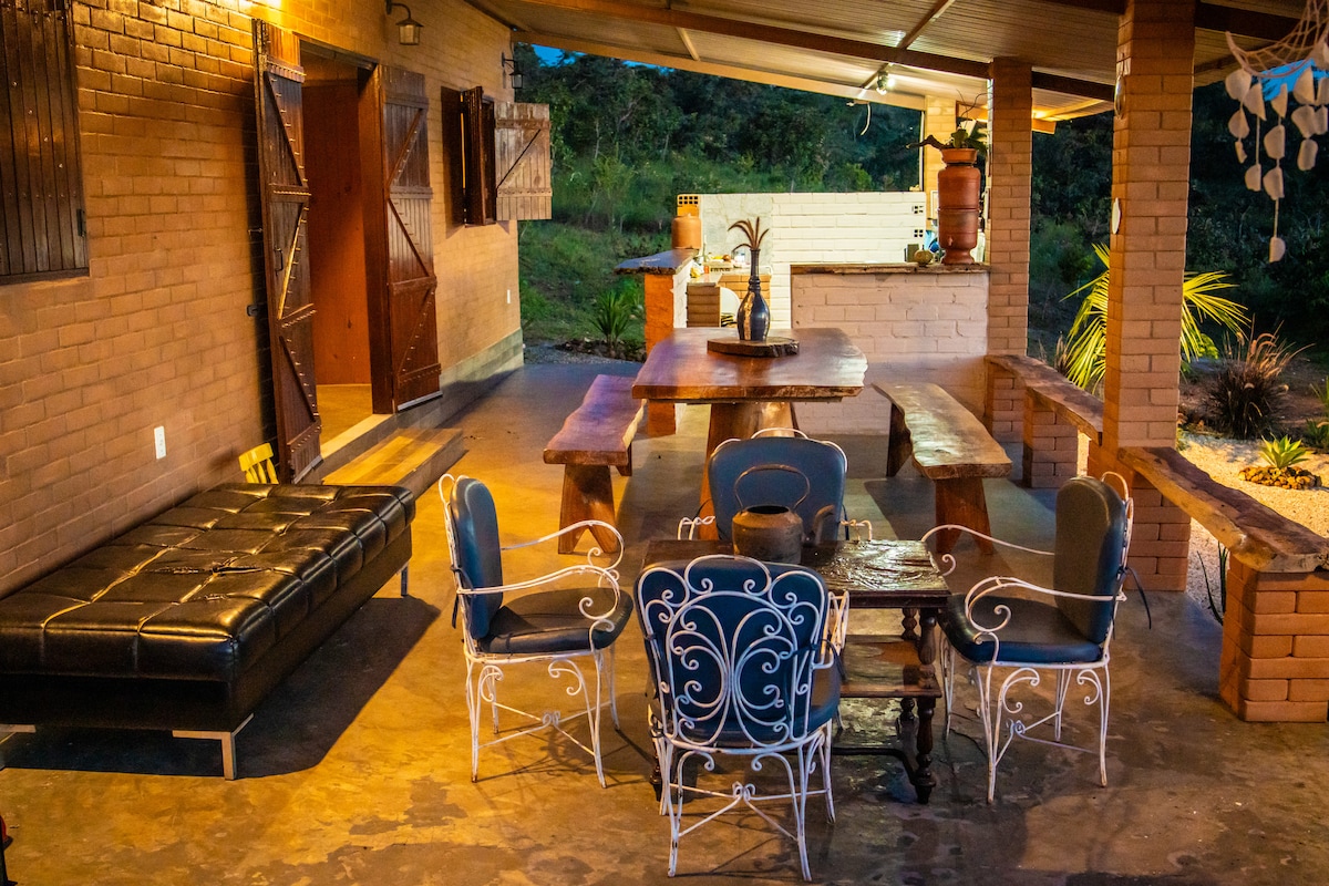 An outdoor seating area features a large wooden table surrounded by various seating options, including a black leather sofa and decorative metal chairs. The space is shaded by an overhang, with natural greenery visible in the background.
