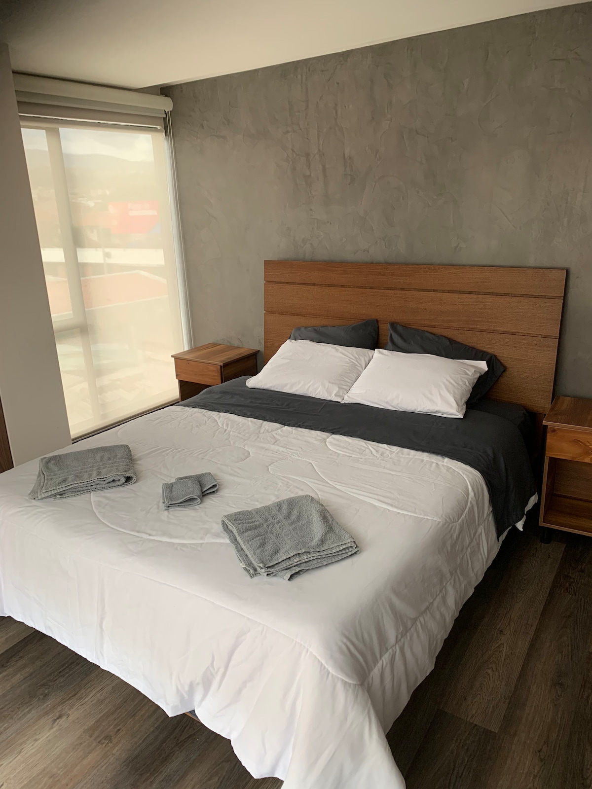 A cozy sleeping area features a spacious bed with crisp white linens and soft gray accents. Two nightstands flank the bed, enhancing the minimalist design. Natural light floods the room through a large window, with a view of the surroundings.