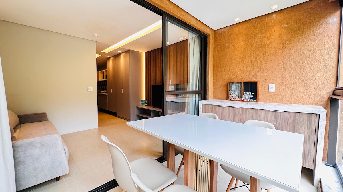 A modern dining area features a sleek white table surrounded by light-colored chairs. A comfortable gray sofa is positioned nearby, with a wooden wall providing warmth to the space. Large glass doors offer natural light and an inviting view of the surroundings.