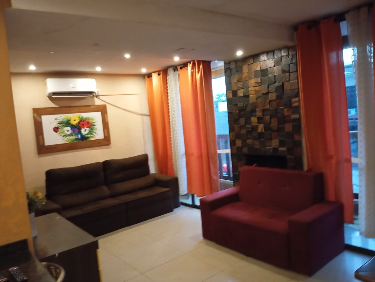 A living area features a cozy layout with two distinct sofas, one in a dark shade and the other in red. Natural light filters through large windows adorned with sheer and colored curtains, illuminating the space. A decorative floral artwork adds a subtle touch.