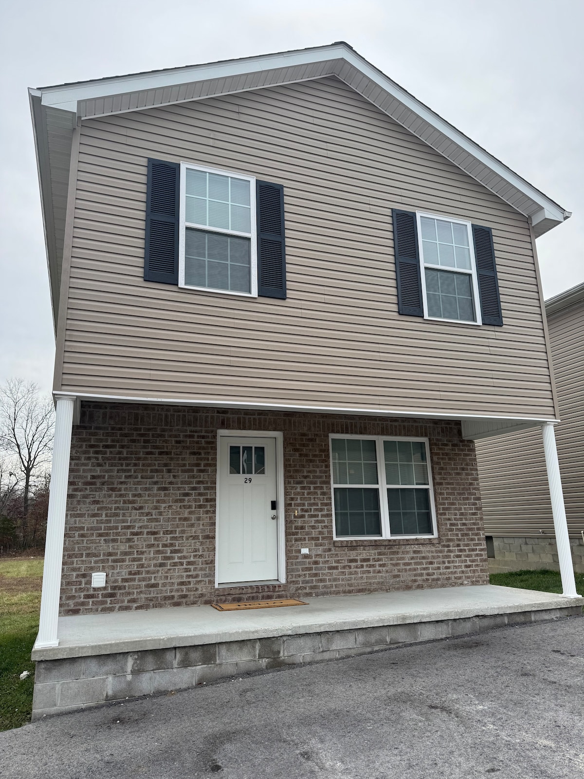 A two-story cottage features a beige exterior with dark shutters on the upper level. A welcoming front porch is supported by white columns. Large windows allow natural light to enter, while a stone foundation adds stability to the overall structure.