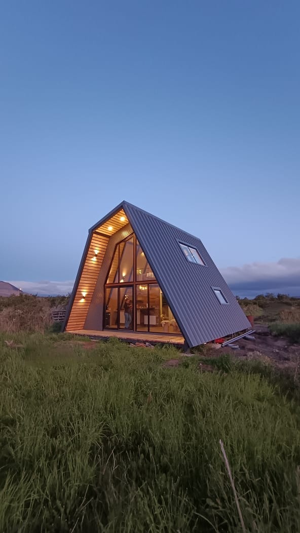 A modern A-frame house stands amidst lush greenery, featuring large glass windows that reflect the evening sky. Soft lighting accentuates the structure’s angles, inviting a sense of calm. The surrounding landscape offers an extensive view of the area, emphasizing the house's connection with nature.