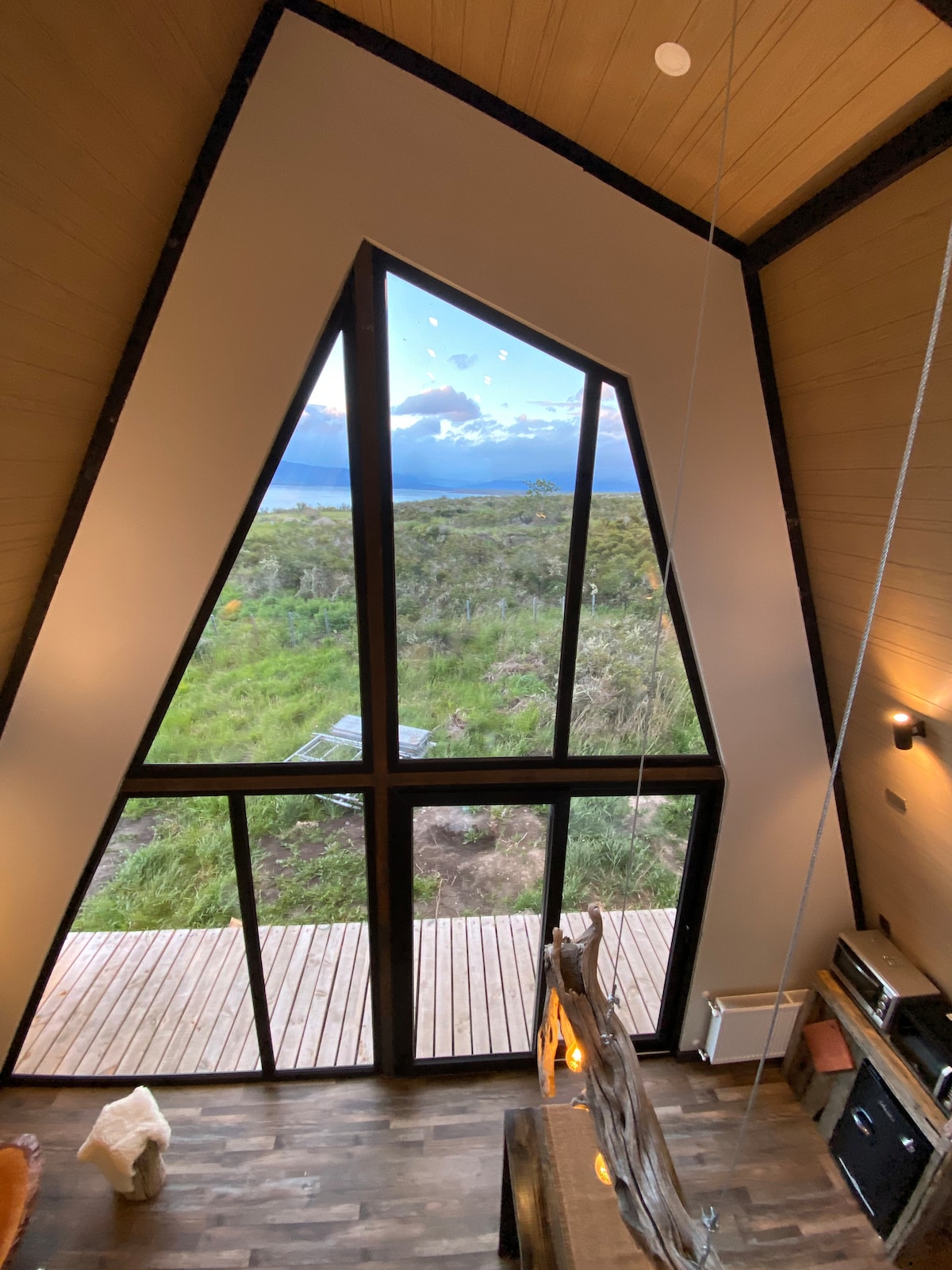 A large triangular window offers a view of the lush landscape outside, featuring greenery and distant mountains. Natural light floods the interior, highlighting the wooden flooring and contemporary design elements, creating a spacious and airy environment.