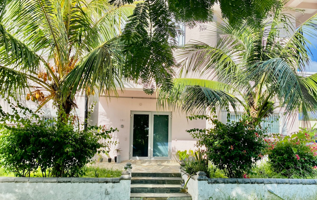 An inviting entrance is framed by lush tropical foliage, featuring tall palm trees and vibrant greenery. A set of stone steps leads to large glass doors, creating a welcoming access point to the building.