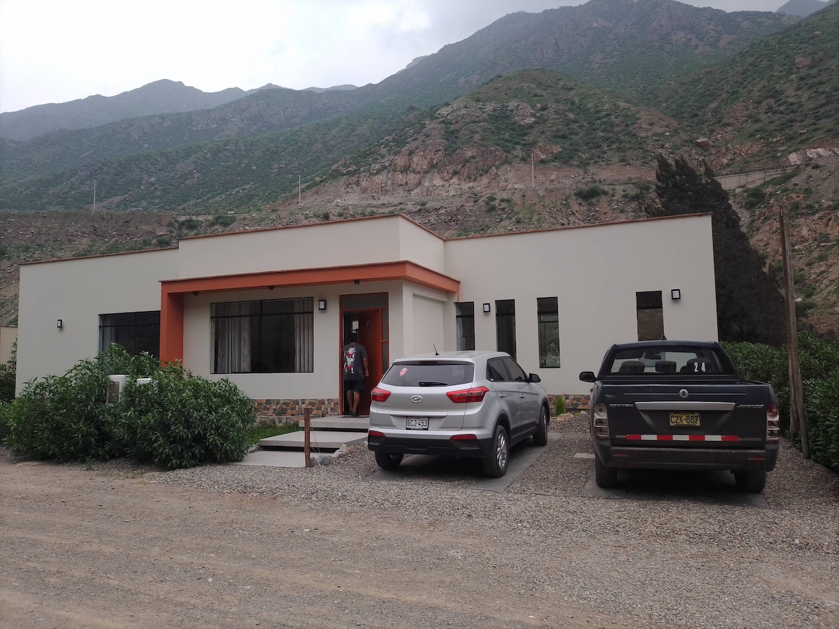A modern single-story house is set against a mountainous backdrop. Large windows allow natural light to fill the space, while the entrance is accented by a covered porch. Two vehicles are parked outside, surrounded by gravel and greenery.
