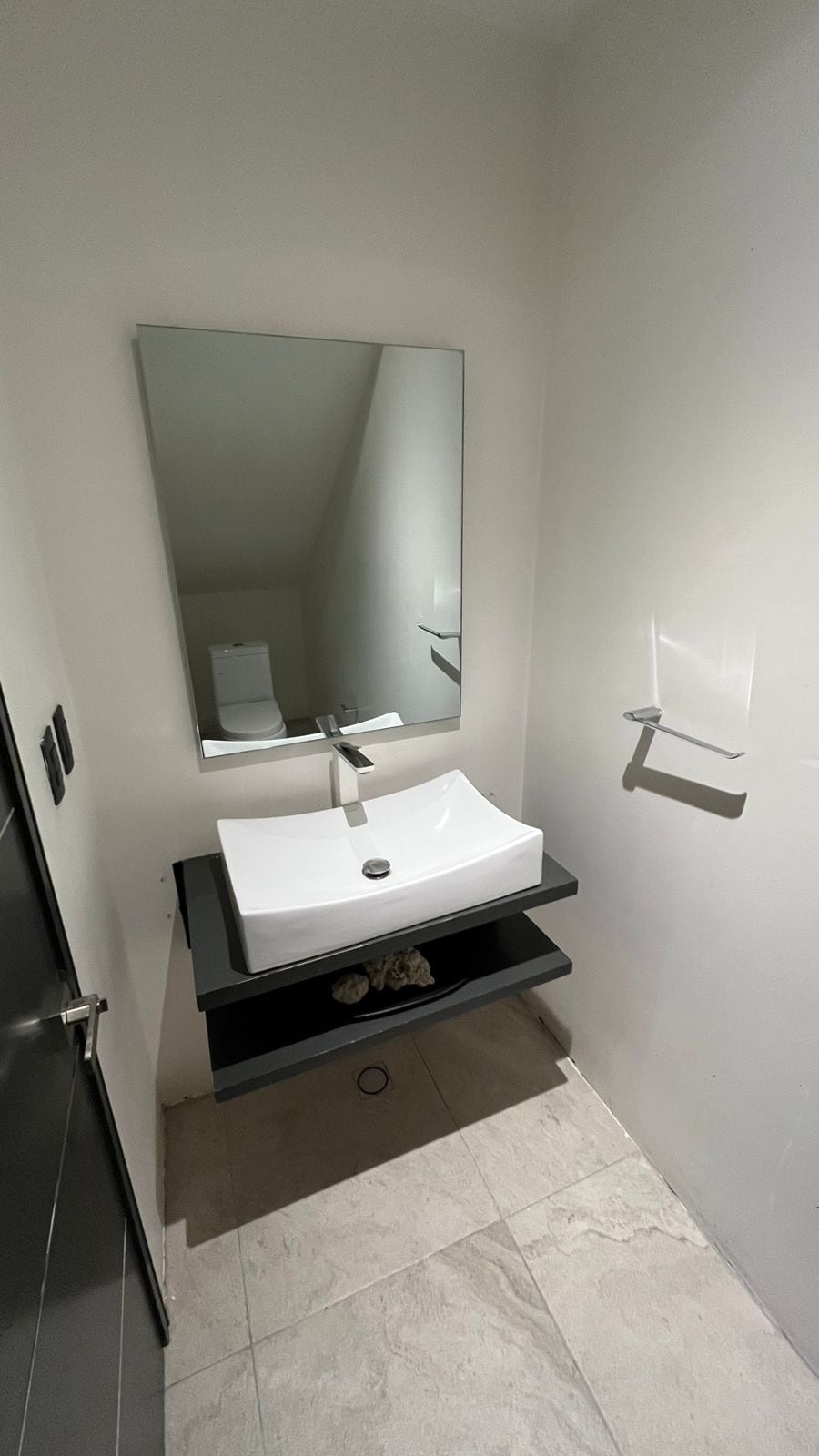 A minimalistic bathroom features a modern pedestal sink with a sleek faucet. A large mirror reflects the well-lit space. Below, a dark shelf holds decorative items. Neutral-colored tiles cover the flooring, contributing to the clean aesthetic.