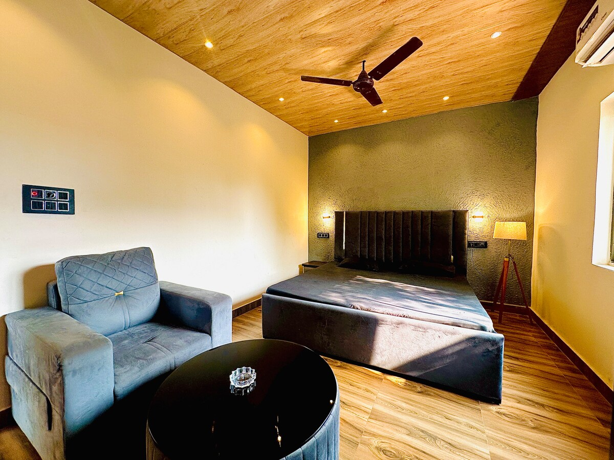 A spacious room features a large bed with a dark upholstered headboard, complemented by a comfortable armchair and a round coffee table. Soft lighting highlights the textured wall, while natural light enters through the nearby window, enhancing the room's serene atmosphere.