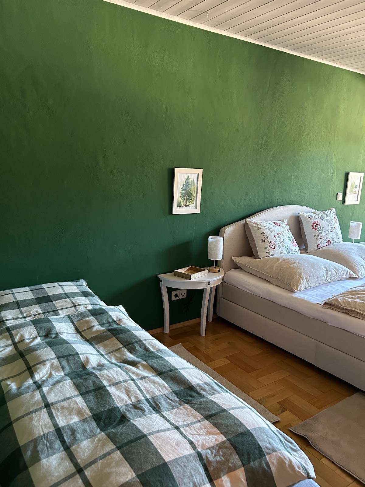 A serene sleeping area is presented, featuring a spacious bed with decorative pillows alongside a single bed adorned with a checkered blanket. Green walls create a calming backdrop, while light wooden flooring adds warmth. Framed artwork is visible on the walls.