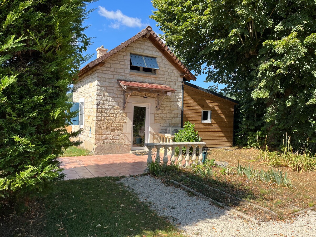 A charming stone house is surrounded by greenery, showcasing natural textures and colors. A welcoming entrance is presented with a small porch, where outdoor seating is visible. Sunlight filters through the trees, offering a serene atmosphere.
