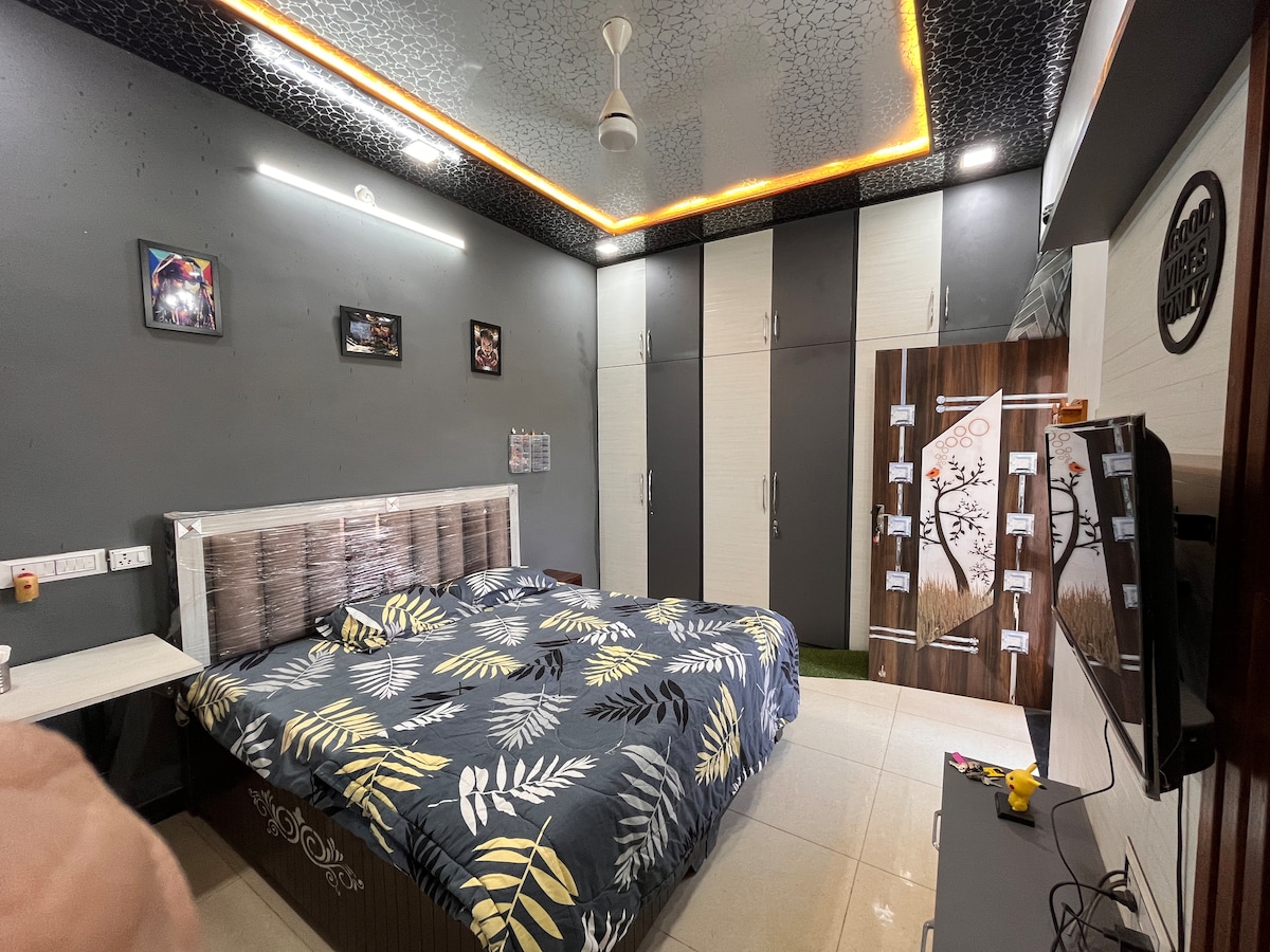 A spacious bedroom features a large bed adorned with a modern leaf-patterned duvet cover. Storage cabinets line the wall, alongside framed artwork. A flat-screen TV is positioned on a stylish stand, and a fan is mounted on the ceiling, providing comfort in the room.