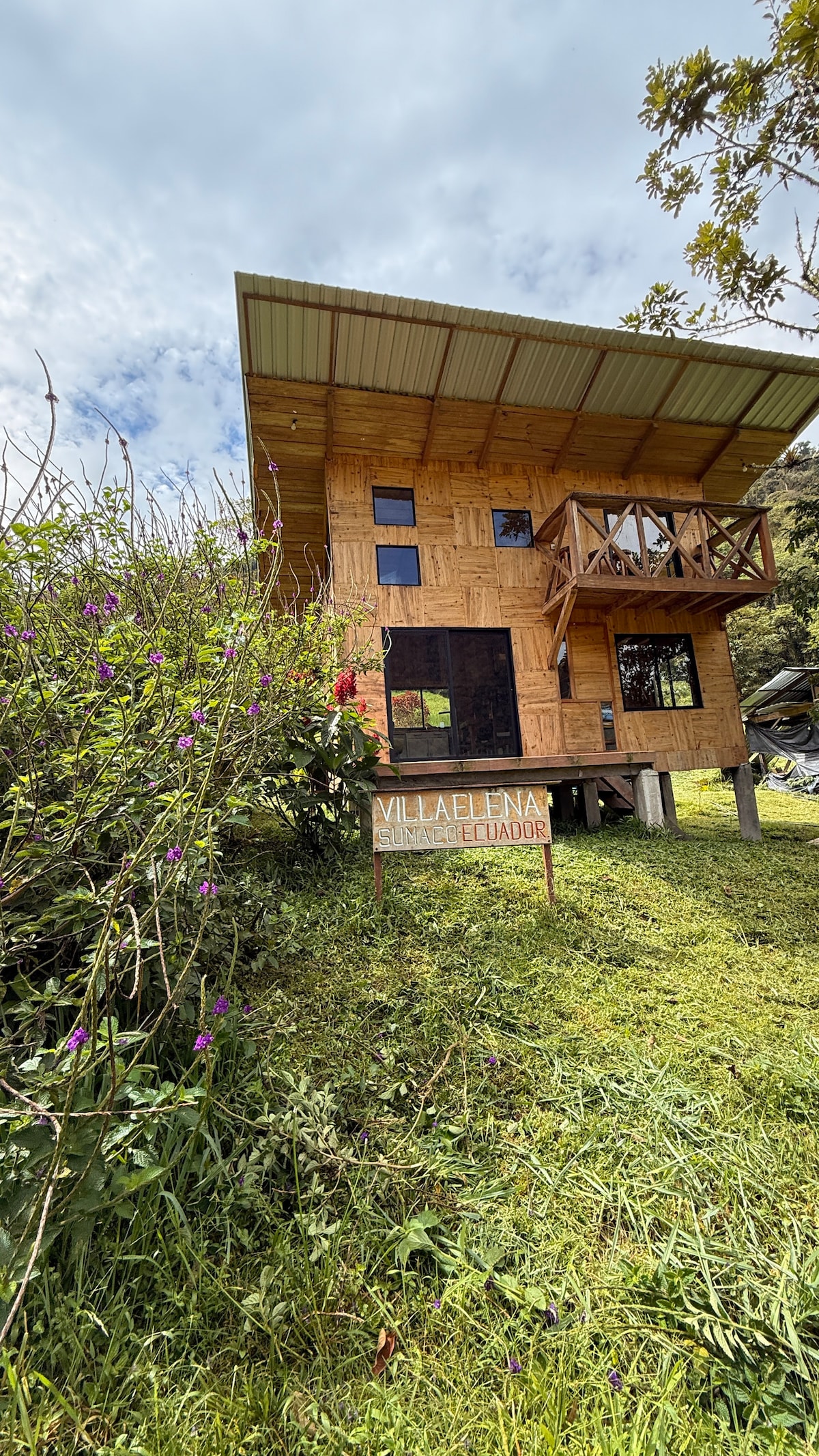A two-story wooden cabin stands amidst lush greenery, featuring a welcoming sign that reads 'Villa Elena Ecuador.' Large windows and a balcony enhance the natural light, while flowering plants and tall grass surround the cabin, creating a serene setting.