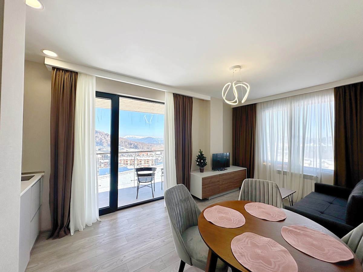 A spacious living area features large glass doors that open to a balcony, allowing light to fill the room. Soft curtains frame the windows. A dining table with round placemats sits beside a comfortable seating area, and a television is positioned on a side cabinet.