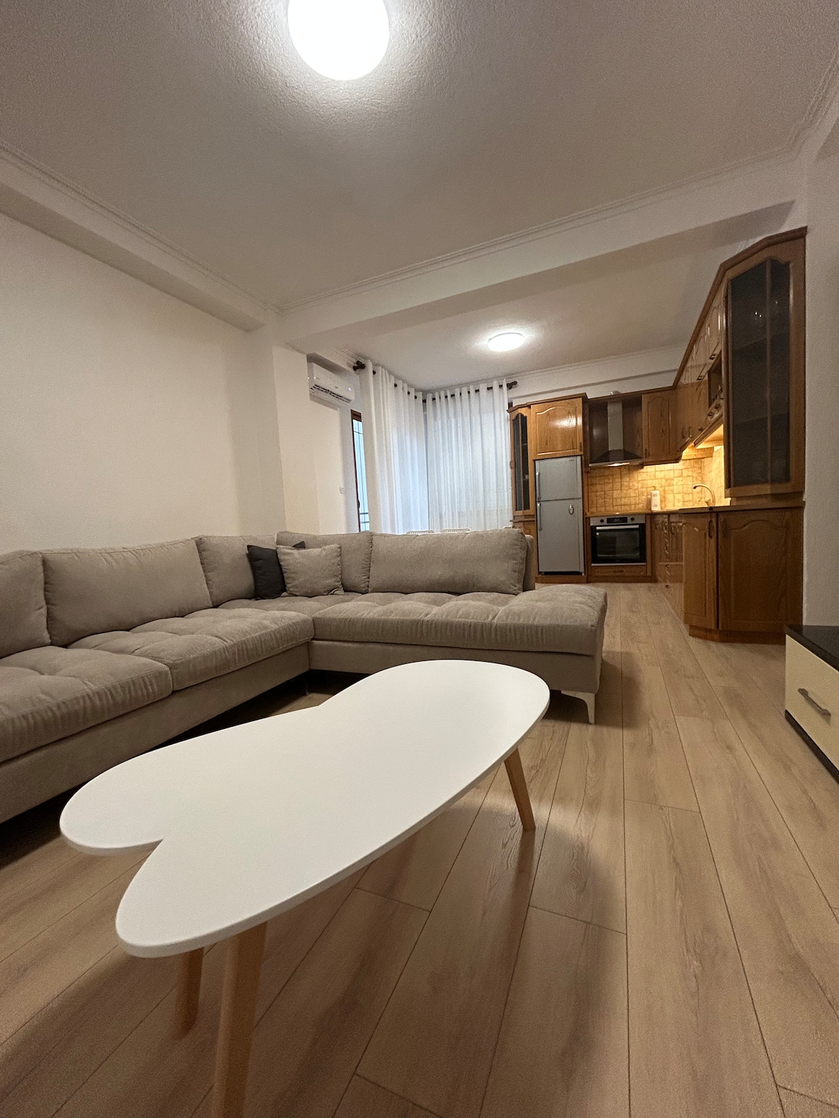 The living area features a spacious light-colored sectional sofa arranged around a heart-shaped coffee table. Natural light filters through sheer curtains, illuminating the nearby kitchen with wooden cabinetry and modern appliances.