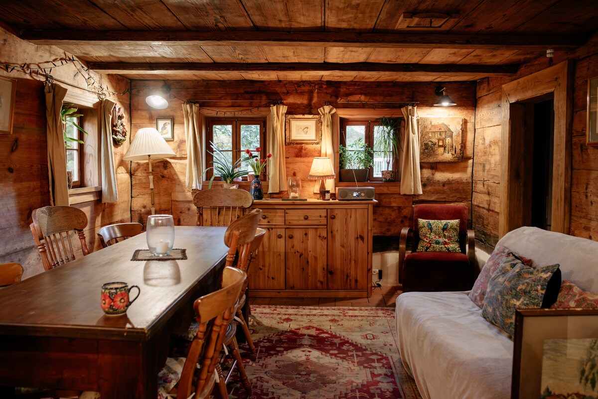 A rustic interior features exposed wooden beams and walls, creating a cozy atmosphere. A long dining table is surrounded by wooden chairs. Soft lighting illuminates a side cabinet and a plush armchair, while natural light filters through multiple windows adorned with curtains.