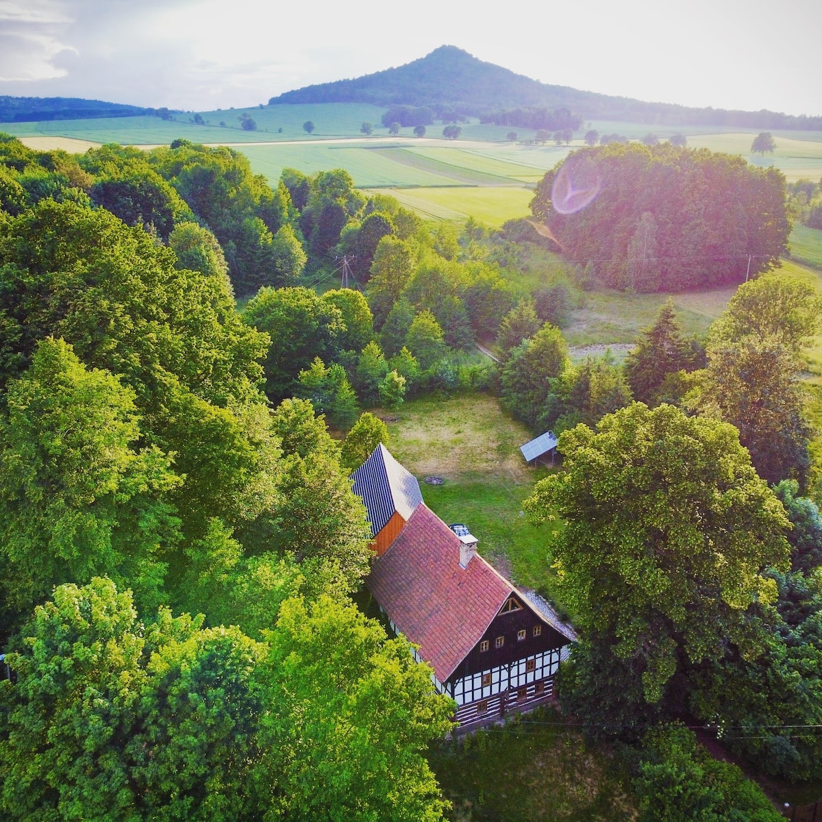An aerial view of a charming house surrounded by lush greenery, with a sloping hill in the background. The landscape features rolling fields and dense trees, creating a serene atmosphere. The house showcases traditional architecture with a colorful roof that complements the natural surroundings.
