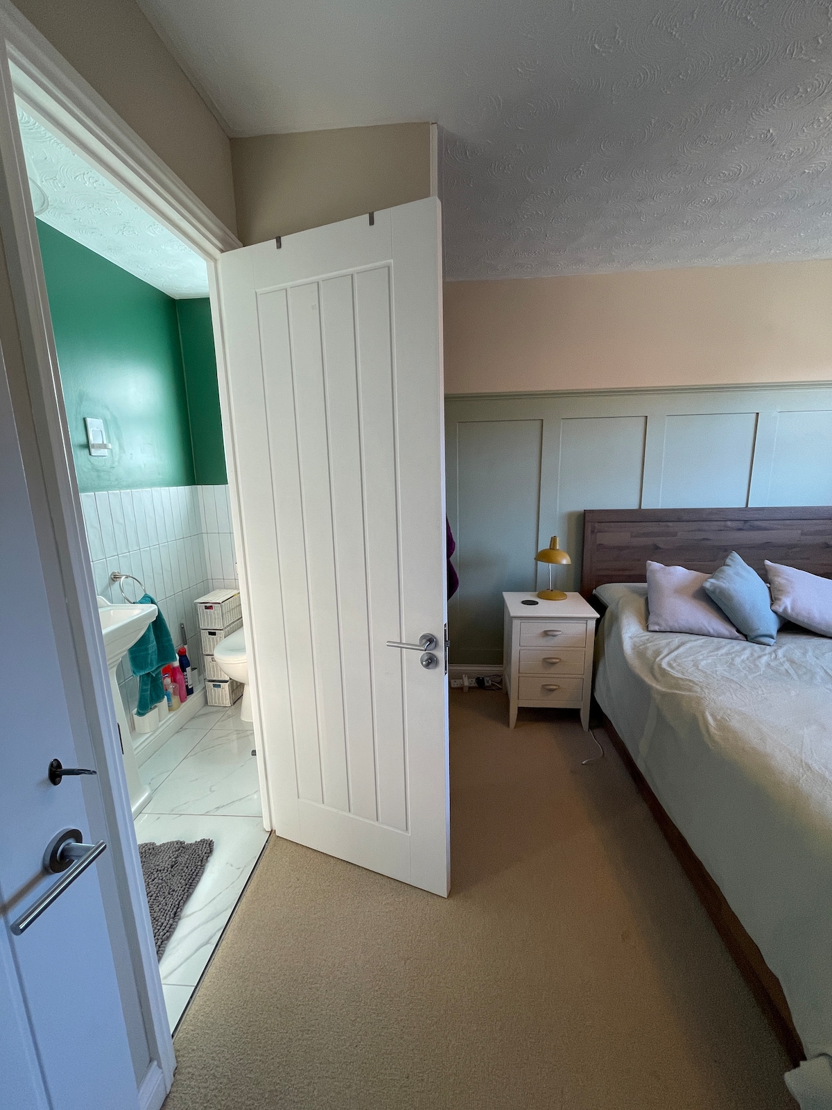 A doorway leads from the bedroom to the bathroom, revealing a well-lit space with a green accent wall. The bedroom features a spacious bed with neutral bedding, complemented by a bedside table topped with a yellow lamp. The area is carpeted and inviting.