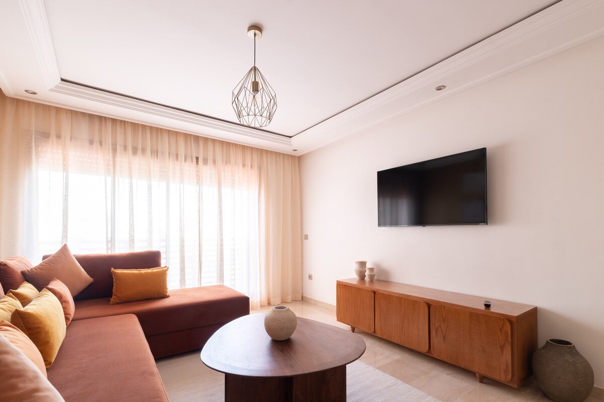 A comfortable living area features a spacious sectional sofa adorned with orange and yellow cushions. A wooden coffee table sits in the center, complemented by a decorative vase. Floor-to-ceiling sheer curtains allow natural light to fill the space, with a flat-screen television mounted on the wall.
