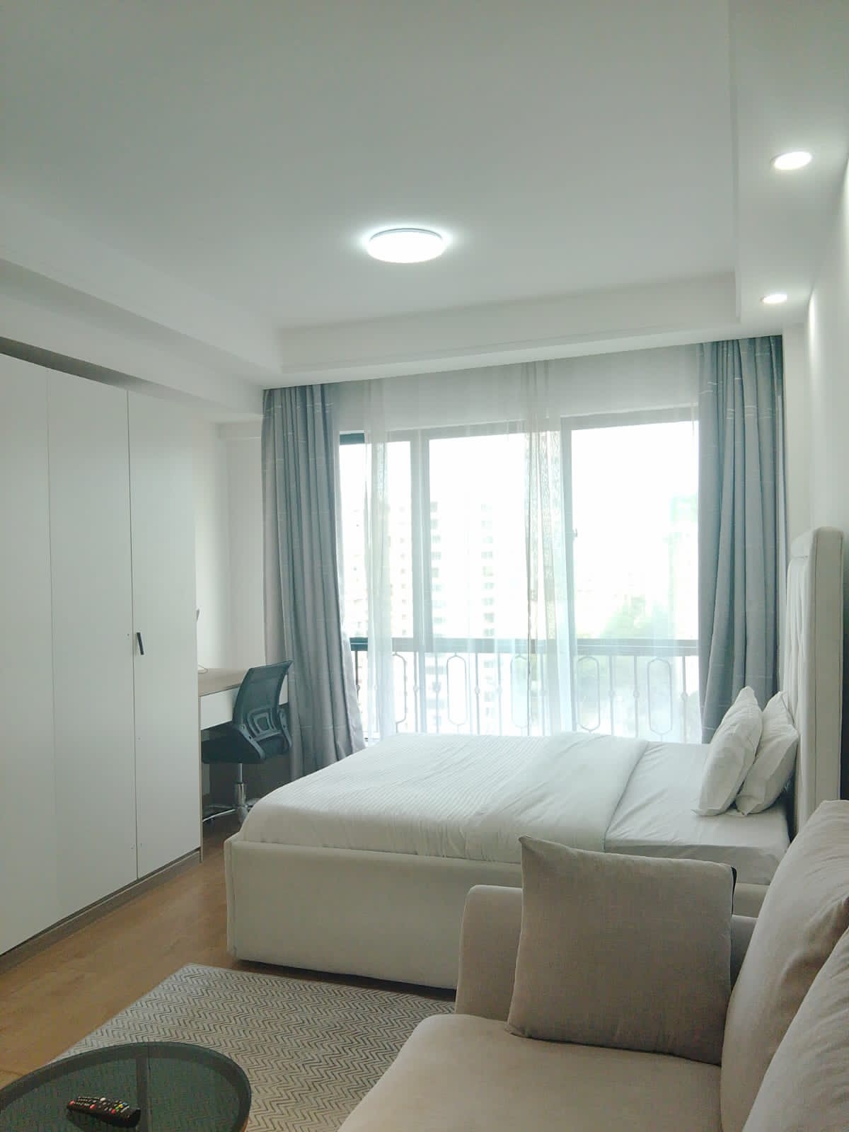 A bright sleeping area features a spacious bed with plush pillows. A sleek, modern desk is positioned near the window, allowing natural light to fill the room. Sheer curtains frame the glass doors that lead to a balcony, enhancing the open feel of the space.