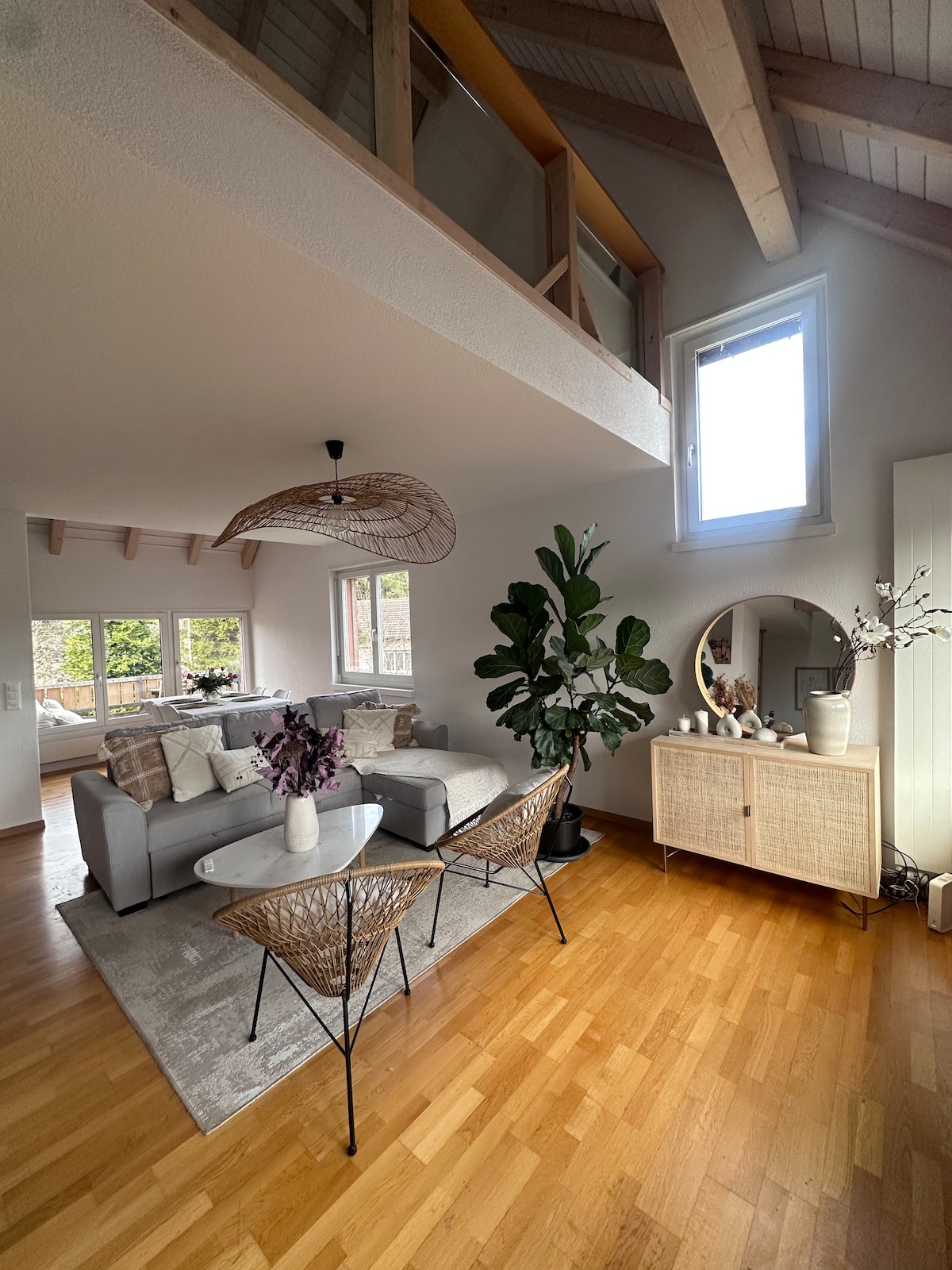 The bright living area features a cozy gray sofa adorned with decorative pillows and a light rug. Two woven chairs are positioned around a small table. Large windows allow ample natural light, while plants add a touch of greenery to the space.