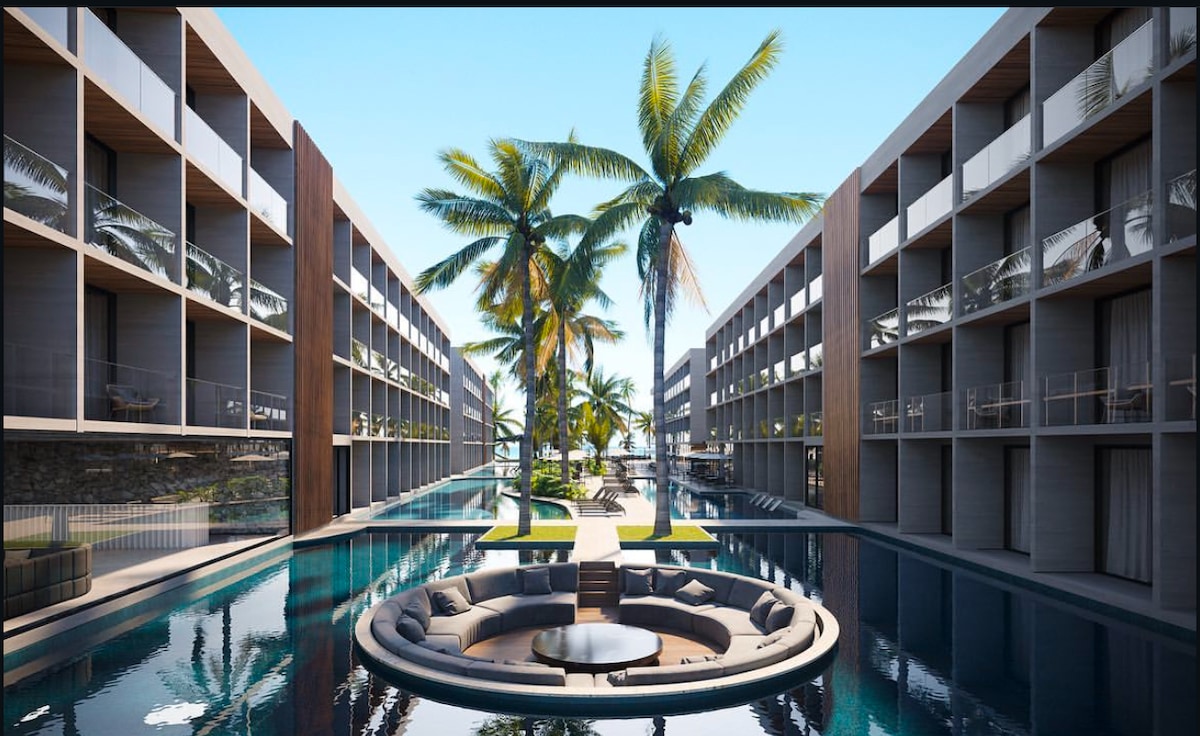 A modern resort layout is depicted, featuring elegant accommodation buildings flanking a serene reflection pool. Central to the space is a circular lounge area surrounded by palm trees, providing a tranquil spot for relaxation and socializing.