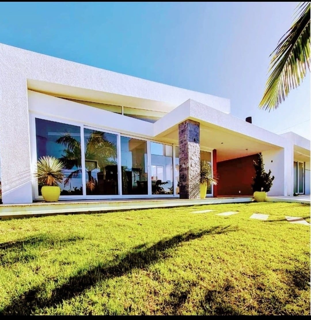 The modern façade of the house features large glass panels and a sleek design, framed by vibrant green grass. Potted plants and a stone accent add to the contemporary aesthetic, while the clear blue sky enhances the inviting atmosphere of the property.