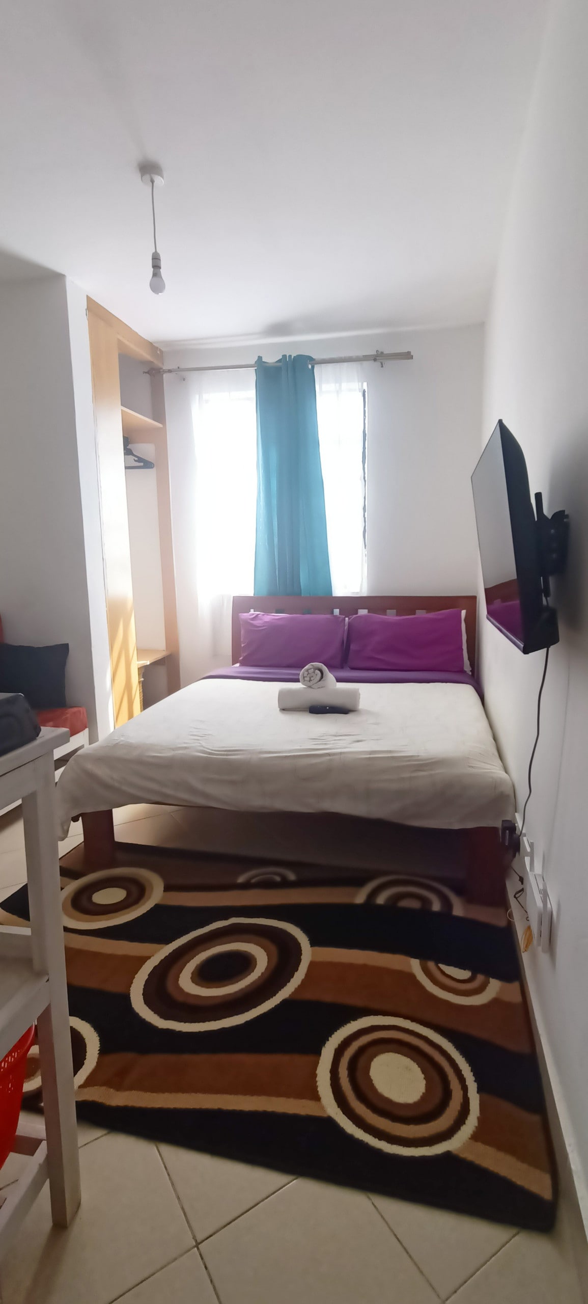 A cozy sleeping area features a spacious bed with purple accent pillows and a neatly rolled towel. A vibrant, circular patterned rug covers the floor, while a window with sheer curtains allows natural light to brighten the room. A wall-mounted television is visible across the bed.