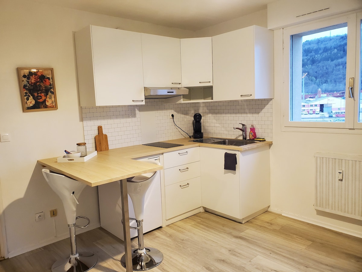 A modern kitchen is displayed, featuring white cabinetry and a wooden countertop. Two bar stools are positioned at the counter. A small appliance is visible alongside a sink and stove. Natural light enters through a window, allowing a view of the surrounding greenery.