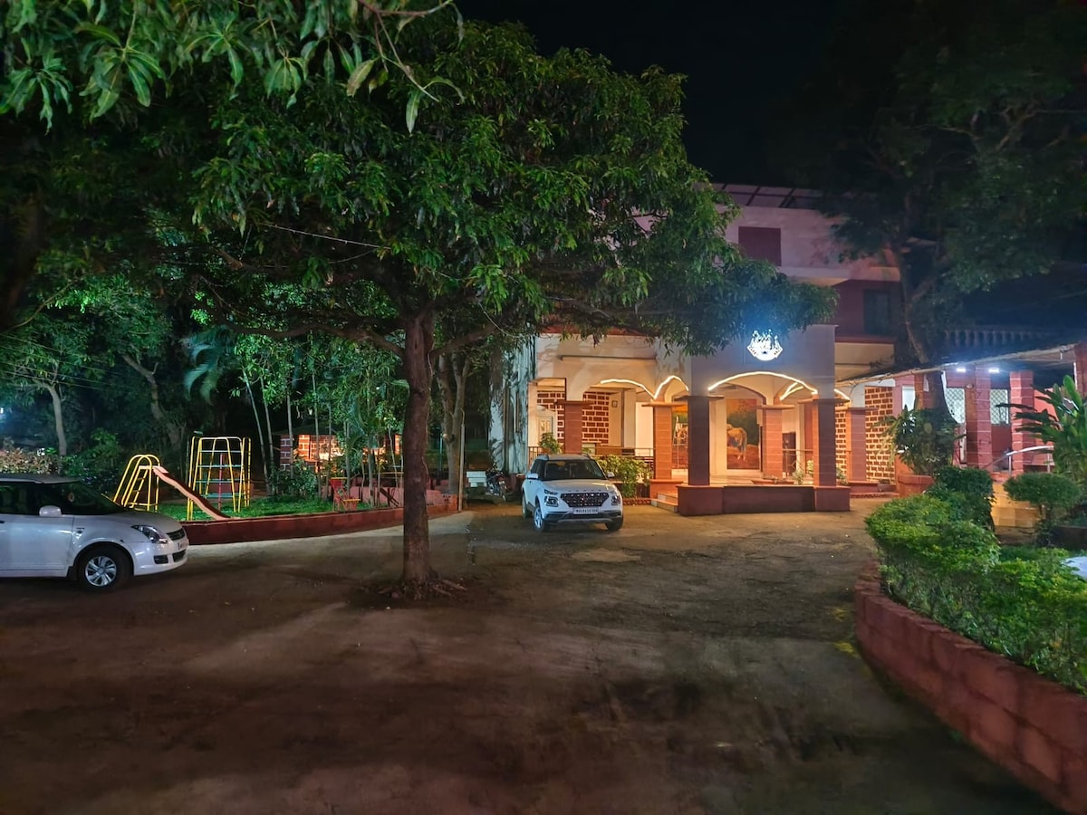 The exterior view of the resort presents a well-lit building surrounded by lush green trees. A parking area is visible, with a parked car positioned nearby. A children's play area is seen in the background, complemented by soft lighting that enhances the evening ambiance.