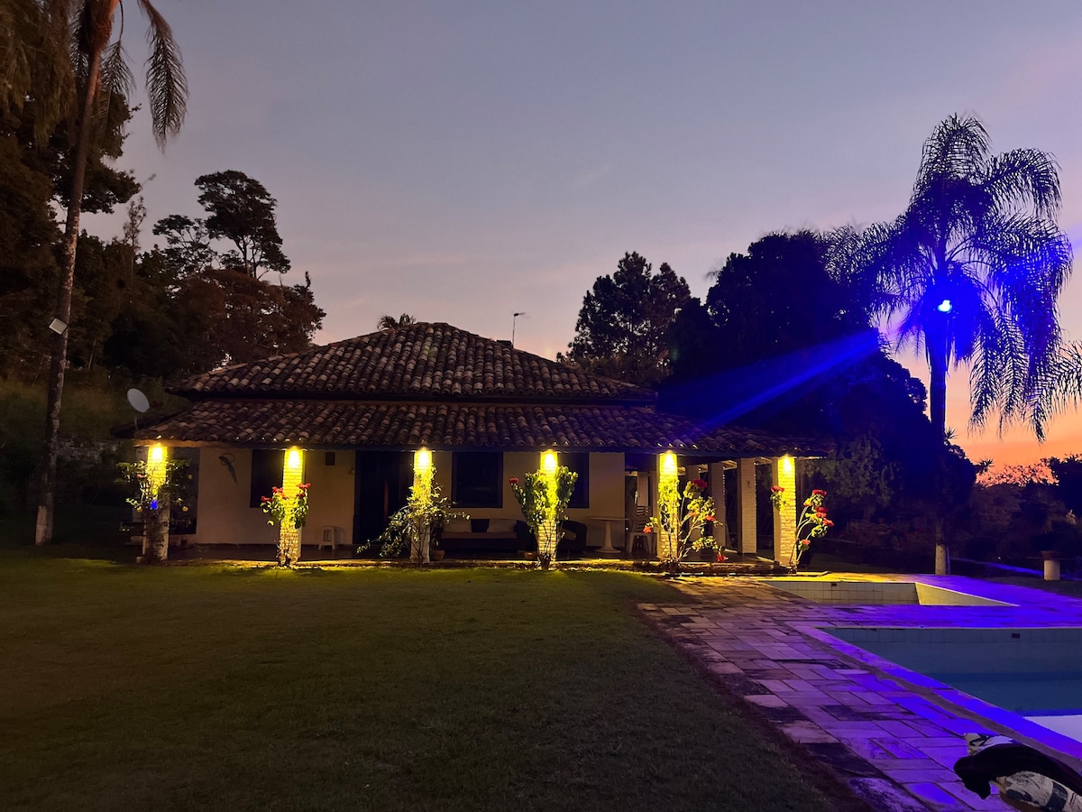 An exterior view of a single-story house is seen at dusk, illuminated by soft lighting. Lush greenery and flowering plants frame the entrance, and a spacious patio area extends towards a pool. The surrounding landscape features trees and a serene atmosphere.
