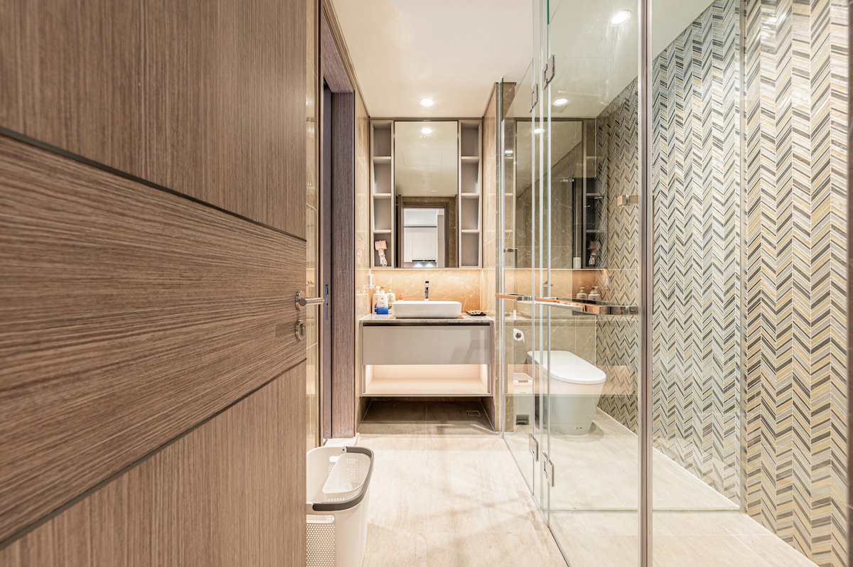 A modern bathroom features sleek wood cabinetry and a glass-enclosed shower. A vanity area with a mirror provides ample space for toiletries. The floor is adorned with neutral tiles, and herringbone-patterned walls add texture to the overall design.