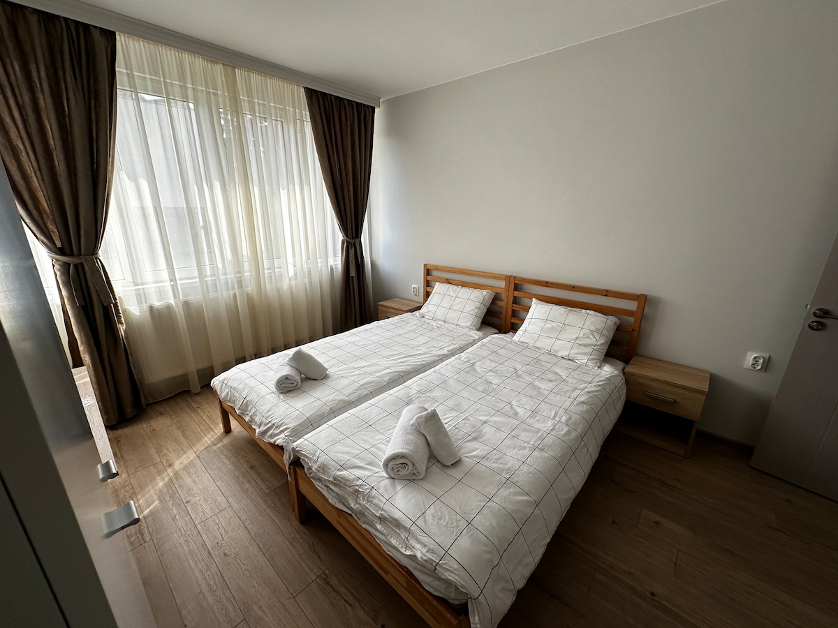 A bright sleeping area features two spacious beds made with white linens, complemented by neatly arranged towels. Floor-to-ceiling windows dressed with sheer curtains allow natural light to fill the room, while light wood flooring enhances the sense of openness.