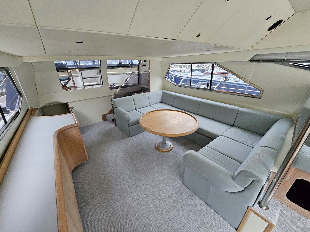 The spacious living area features a circular wooden table surrounded by a curved sectional sofa in soft blue upholstery. Large windows offer views of the marina, allowing natural light to fill the space. The carpeted floor adds comfort to this inviting area.