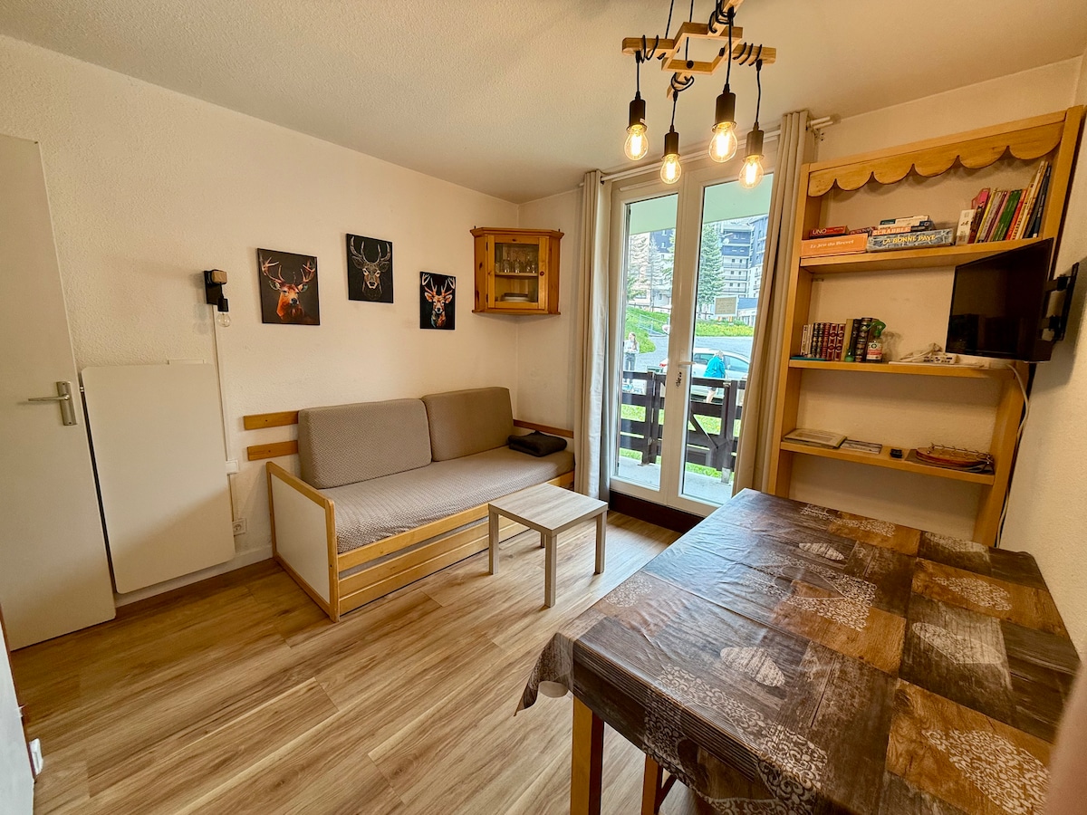A living area is shown, featuring a light-colored sofa and a wooden dining table. Large glass doors open to a balcony, allowing natural light to fill the space. On the walls, framed art and shelves with books add a personal touch to the room.