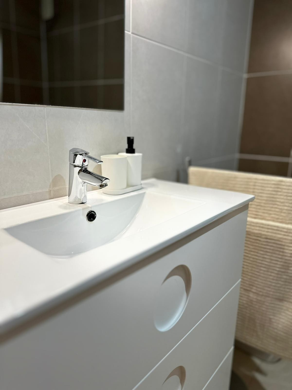 A modern bathroom features a sleek white sink with a polished chrome faucet. Minimalist storage with smooth surfaces is visible alongside a neutral-colored towel, contributing to a clean and uncluttered appearance.