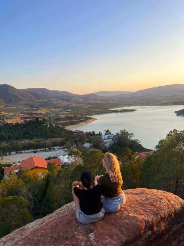 A scenic view of a valley is displayed, featuring lush greenery and a serene body of water. Two individuals are seated on a rock, facing outward toward the landscape, illuminated by the soft glow of sunset. The horizon showcases distant mountains and a clear sky.