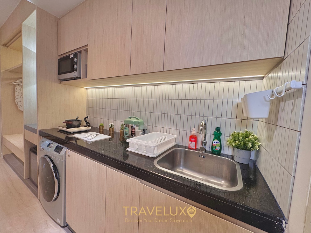The kitchen area features modern cabinetry in light wood tones, with a sleek black countertop. Stainless steel appliances, including a microwave and washing machine, are visible. A variety of utensils and dishware are neatly arranged, along with a small plant adding a touch of greenery.