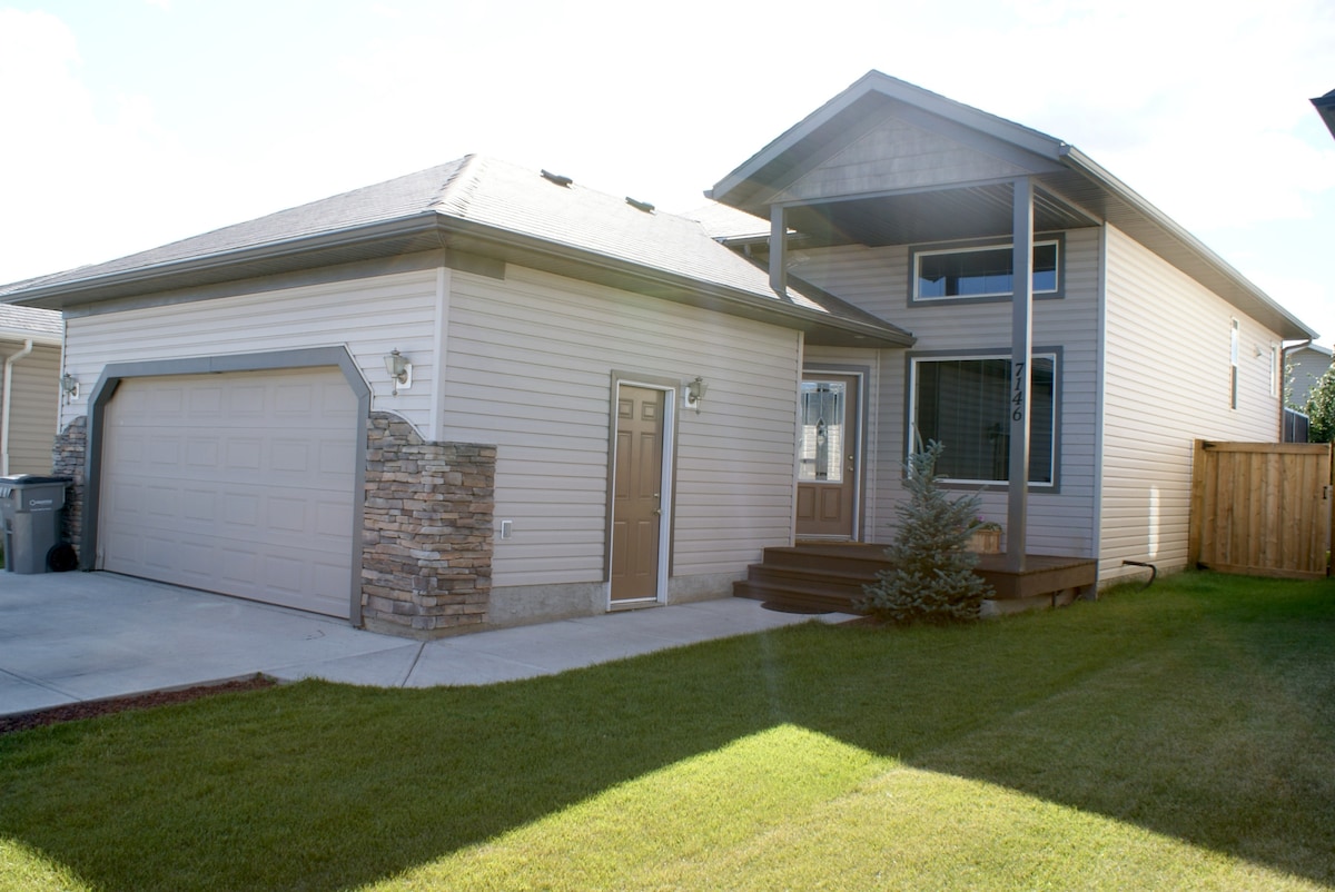 A modern home exterior features a light-colored facade with stone accents. A spacious double car garage is visible alongside a welcoming entry with steps leading to the front door. The yard is well-maintained, showcasing green grass and a nearby fence.