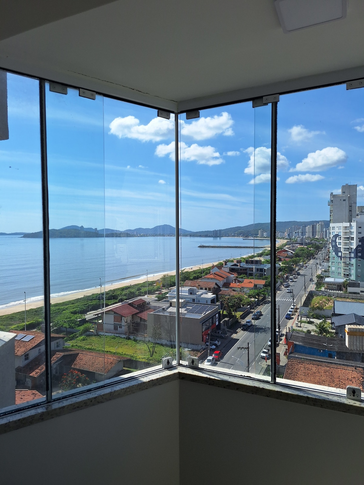 A panoramic view of the coastline is visible through large glass panels. The beach stretches along the water's edge, while distant mountains and city buildings can be seen in the background. Clear skies and soft clouds contribute to the serene atmosphere.
