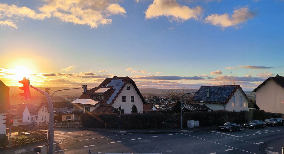 A scenic view at sunset showcases the rooftops of residential homes with varied architectural styles. Solar panels are visible on one roof, and a serene glow envelops the landscape as the day transitions to evening. Traffic signals and vehicles are also present, contributing to the urban setting.