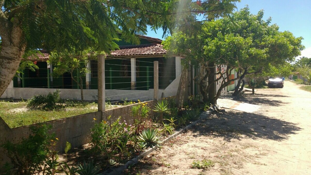 The exterior of a spacious house is surrounded by lush greenery and flowering plants. A sandy path leads to the entrance, framed by trees and shrubs. The house features a traditional roof and multiple windows, allowing natural light to fill the interior.