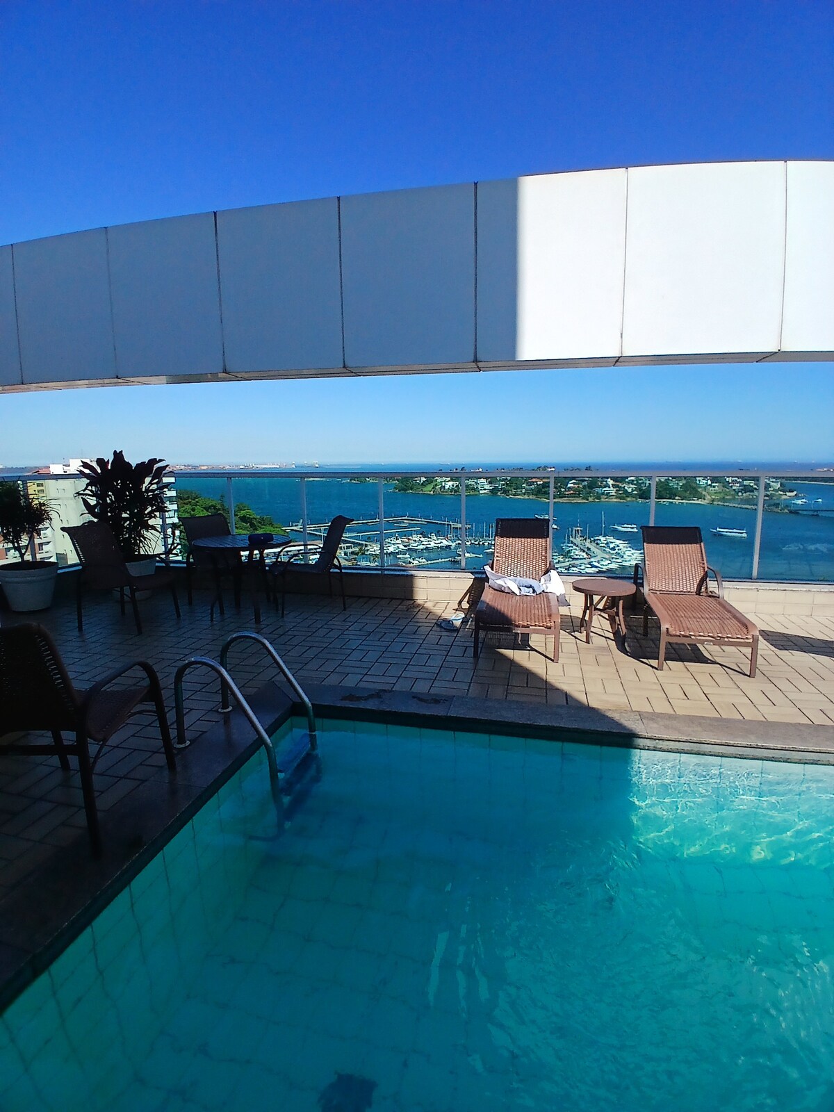A rooftop pool is visible, surrounded by lounge chairs. The water appears clear and inviting, with a panoramic view of the coastline in the background. Sunlight reflects off the surface, and a terrace area features potted plants, enhancing the relaxing atmosphere.