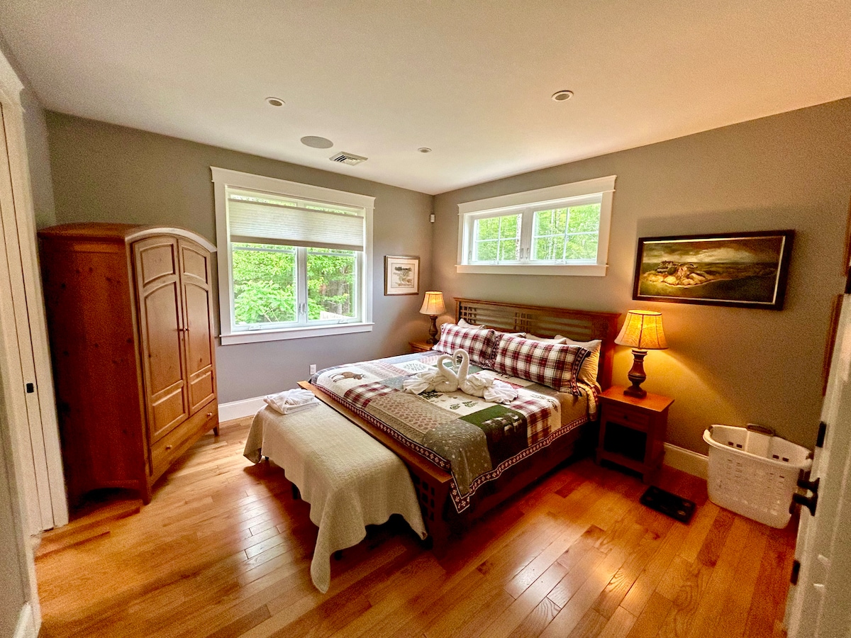 A cozy primary bedroom features a King bed adorned with layered bedding and decorative pillows. Two bedside lamps provide soft light, and a wooden wardrobe stands against the wall. Large windows allow natural light to fill the space, complemented by light-colored walls and hardwood flooring.