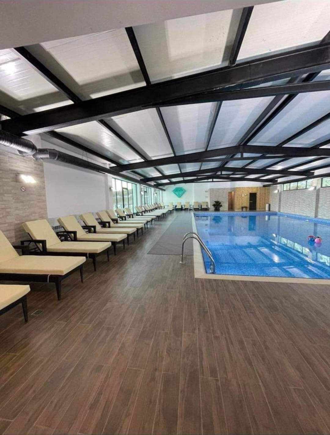 An indoor swimming pool area features a clean and modern design with large windows allowing natural light. Several lounge chairs are arranged along the pool's edge, offering a place to relax. The pool is well-maintained, with clear blue water and a gentle slope.