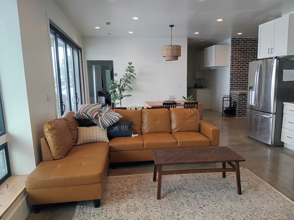 A spacious living area features a warm-colored sectional sofa adorned with decorative pillows. A wooden coffee table sits atop a stylish area rug, with large windows allowing natural light to fill the space. The open layout leads to a modern kitchen in the background.