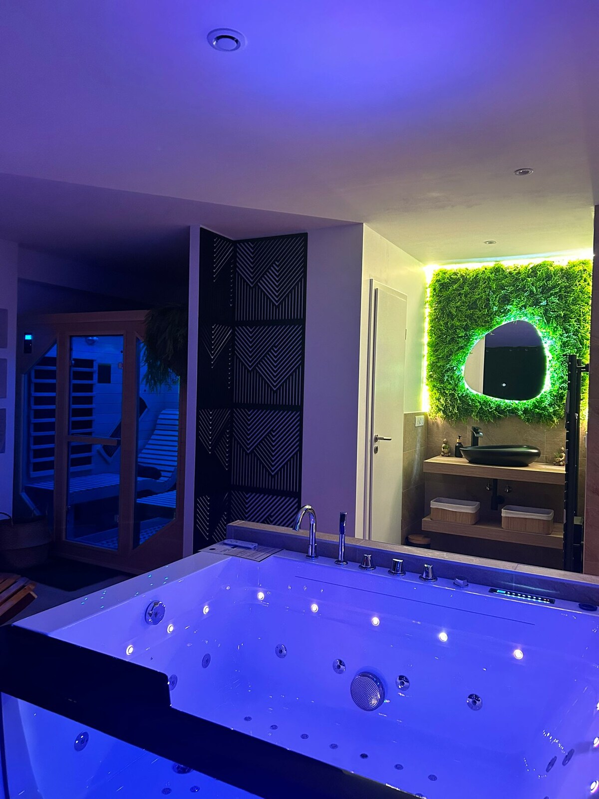A modern wellness area features a large jacuzzi with soft blue lighting, inviting relaxation. A wooden sauna is visible through an open doorway, while a vibrant green moss wall with a circular mirror adds a touch of nature to the space.